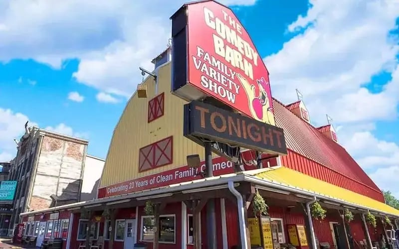 View of the Comedy Barn Theater in Pigeon Forge with red and yellow barn building and marquee sign against blue sky.