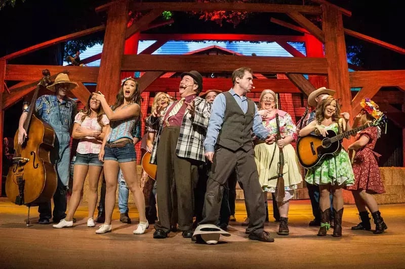 Cast of Hatfield & McCoy Dinner Feud performing music on rustic stage with guitars, fiddle and double bass under wooden beams