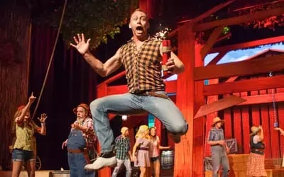 Performer jumps on the Hatfield & McCoy Dinner Feud stage with arms up while other cast members watch near red wooden beams.