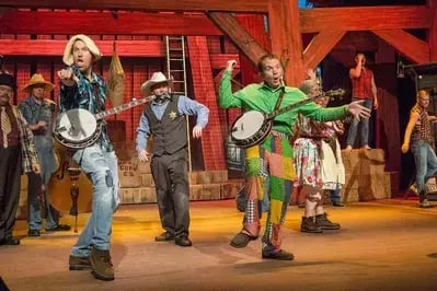 Performers on stage at Hatfield & McCoy Dinner Feud playing banjo and fiddle with rustic barn set and comedic antics
