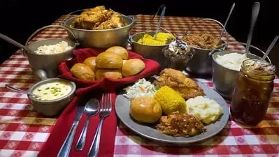 Plate of fried chicken, corn, mashed potatoes, ribs and vegetables on a red checkered table with side dishes and iced tea