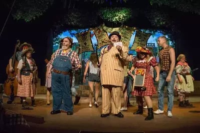 Cast of Hatfield & McCoy Dinner Feud on stage in rustic costumes as man in striped suit addresses group under forest backdrop